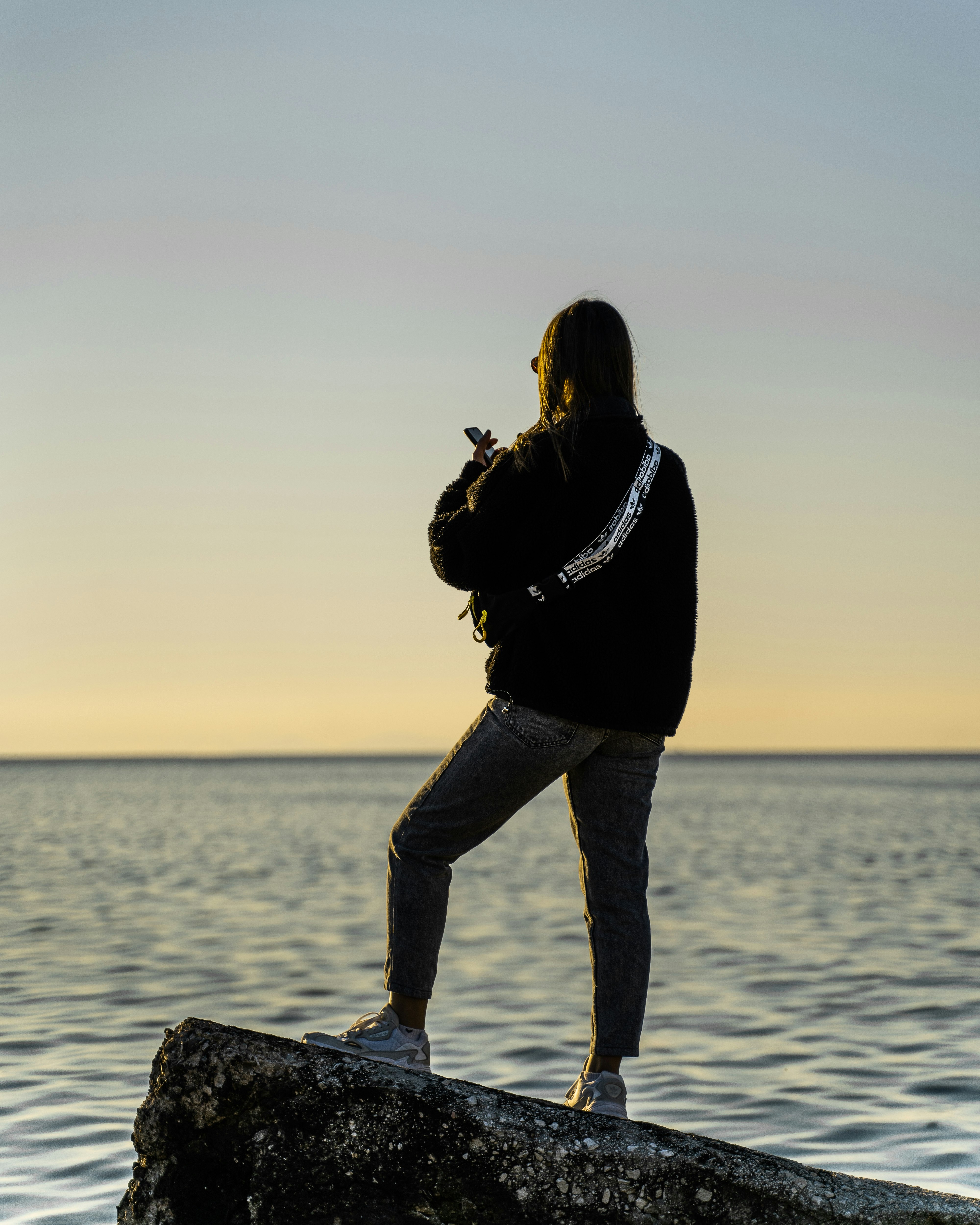 woman in black jacket and blue denim jeans standing on rock near body of water during